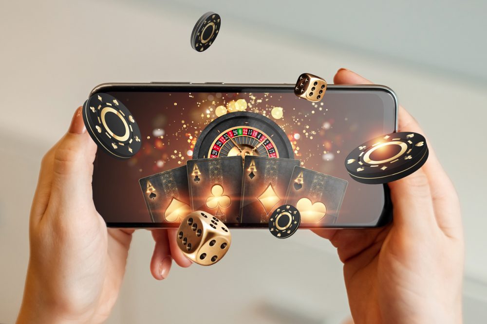 Creative background, online casino, in a man's hand a smartphone with playing cards, roulette and chips, black-gold background. Internet gambling concept. Copy space